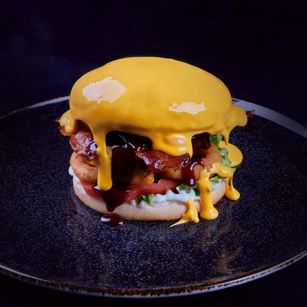 Burger mit Dry Aged Beef Patty, Cheddar, knusprigem Bacon, Zwiebelringen, Röstzwiebeln, Tomate, Lollo bionda, Trüffelmayo, BBQ Dip mit Biergeschmack und Chili Cheese Dip